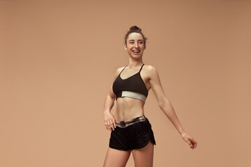 Fototapeta na wymiar Portrait of young, smiling girl with sportive, fit body posing in sportswear against light brown studio background. Body-positivity. Concept of sportive lifestyle, beauty, body care, fitness, health