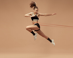 Fototapeta na wymiar Dynamic image of sportive, fit, strong young girl training, in motion, jumping with fitness string against light brown studio background. Sportive lifestyle, beauty, body care, fitness, health concept