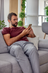 Relaxed young handsome bearded man sitting on sofa and using cellphone