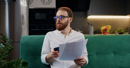 Man beard managing finances, using calculator sitting on sofa in kitchen. Finances concept. Focused man analyzing data at home.