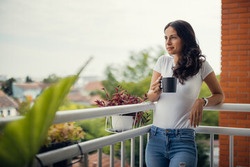 A beautiful young woman standing on the balcony holding a cup of coffee.