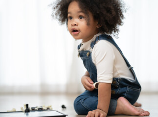 Little kid girl play chess toy board at home with fun. Portrait of adorable multiracial daughter sitting on floor enjoy playing piece of chess isolated on white background. Joyful leisure childhood.