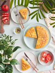 Delicious Spanish potato omelette served in portions on the table. Top view of the table with salad tomatoes and decoration of green leafy plants.