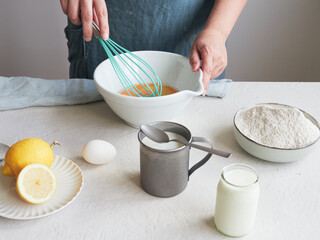 Woman beating eggs with a whisk in a bowl. Ingredients to prepare a dessert or pastry recipe. Flour, milk and lemon on the table. - obrazy, fototapety, plakaty