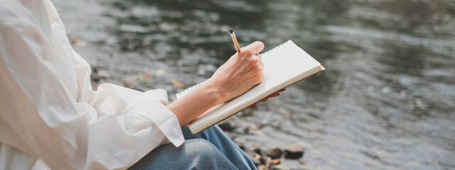 Wide screen of Female's hand is writing or sketching an idea while chilling among nature. Concept of vacation, weekend activity. Write diary in notebook. Natural healing relaxing drawing. Background - obrazy, fototapety, plakaty