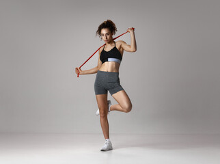 Strenght and endurance. Portrait of young girl in sportswear with slim body posing with fitness expanders against gray studio background. Concept of sportive lifestyle, beauty, body, fitness, health