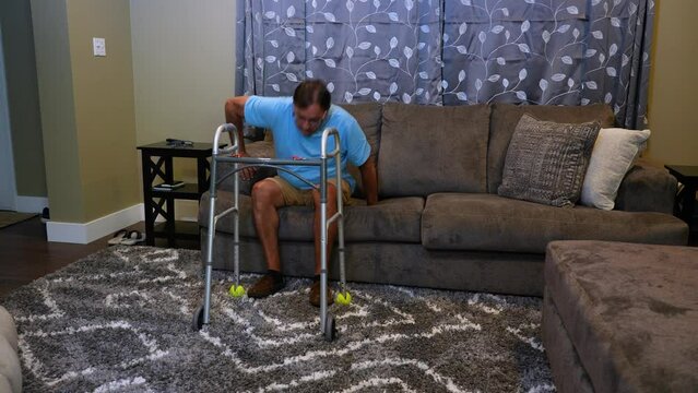 A wide shot of a man in his living room using a walker to get off the sofa and walk to the hallway.  	
