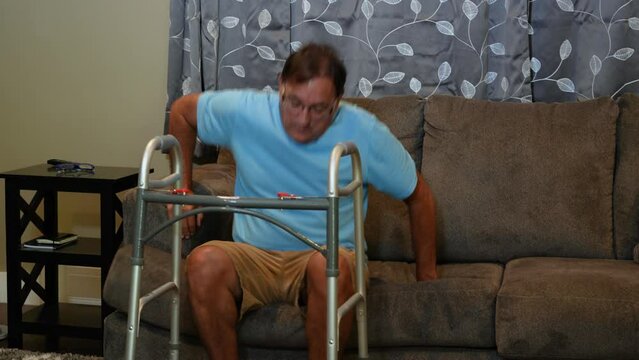 A medium shot of a man in his living room using a walker to get off the sofa and walk to the hallway.  	