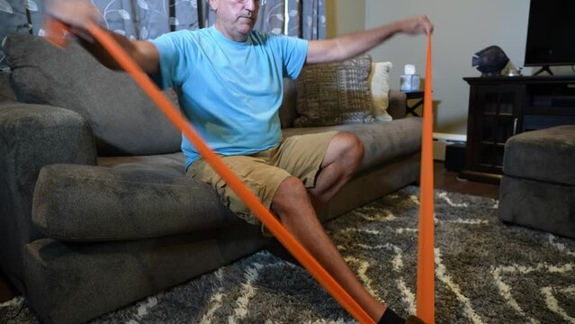 A wide view of a man using an orange rubber exercise band while sitting on his couch in the living room.  	