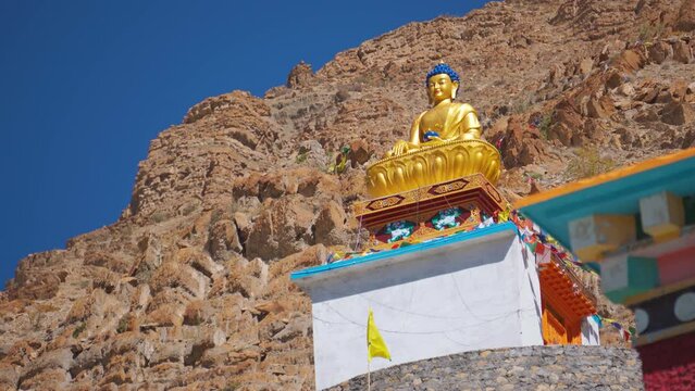 4k view of Golden Buddha statue with prayer flags waving in wind against mountains at Sakaya Tangyud monastery at Kaza, Spiti Valley, India. Buddha statue during sunny summer day. Religious background