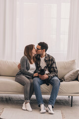 Happy couple hugging and kissing at home sitting on the couch.A French Bulldog puppy is on their...