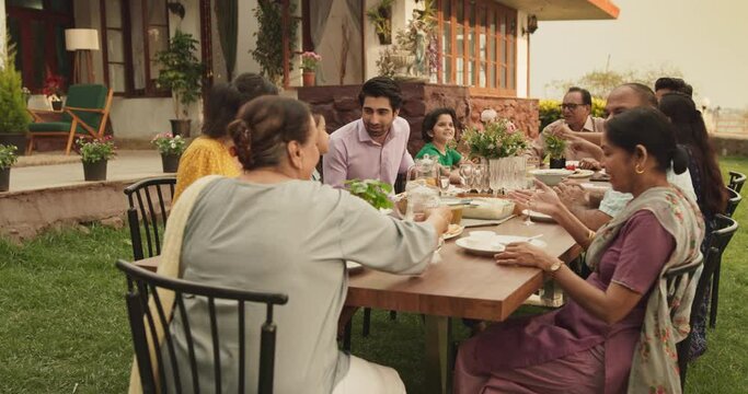 Indian Family Gathering Portrait: Family and Friends Celebrating Outside at Home. Diverse Group of Children, Adults and Seniors Sitting at a Table, Having Fun Conversations. Eating Traditional Food