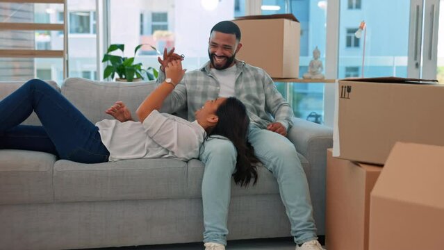 Happy couple, keys and relax on couch in new home with game, playing or bond with love for goal. Man, woman and happiness for young homeowner, achievement and comic with boxes, package or property