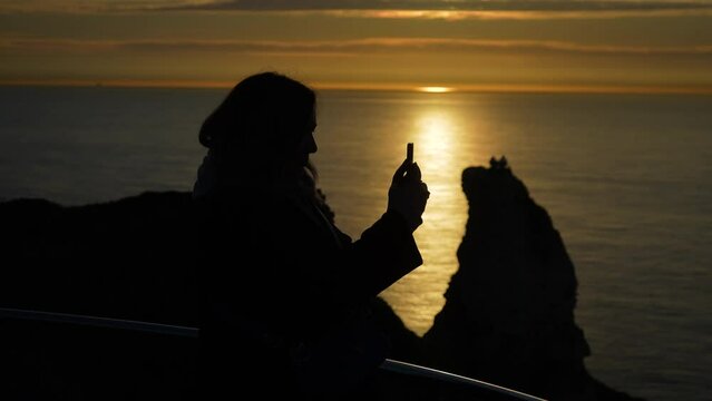 Dark silhouettes of unrecognizable female photographer enjoying sunset sea horizon standing on high mountain, taking photo on smartphone. Young woman watching panoramic evening marine landscape alone