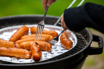 The hands of a person holding two forks change the position of the sausage on the grid of the garden grill. Photo taken in natural lighting conditions.