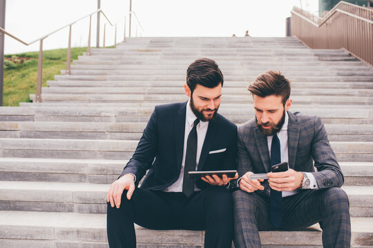 two businessman sitting outdoors staircase using sharing tablet