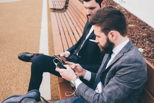 Two young businessman sitting on a bench using smart phone