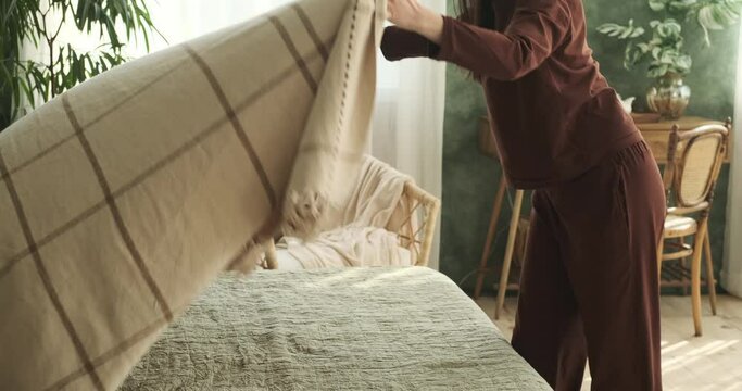 A Caucasian woman, dressed in cozy pajamas, enters tranquil bedroom. She approaches the inviting bed and begins to meticulously arrange the sheets and fluff the pillows.
