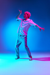 Obraz premium Full-length portrait of mature man in white shirt and checkered pants dancing against blue studio background in pink neon light. Concept of human emotions, lifestyle, youth, facial expression