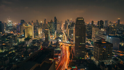 Night cityscape skyline panorama of modern Bangkok city and transport light trails on highway. Illuminated skyscrapers and moving vehicles lights on the road. cinematic color. High-angle view