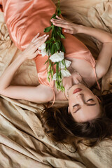 Obraz na płótnie Canvas top view of young woman in pink silk slip dress lying on linen fabric with closed eyes and holding white flowers on mottled beige background, sensuality, sophistication, elegance, eustoma