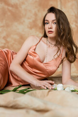 Obraz na płótnie Canvas pretty young woman in pink silk slip dress lying on blurred linen fabric and looking at camera near white flowers on mottled beige background, sensuality, sophistication, elegance, eustoma