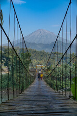 People and motorbike riders cross a wooden suspension bridge over the river with beautiful mountain views. Rural scene of Indonesian countryside