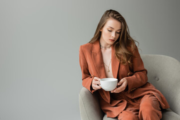 Fashionable young woman in terracotta jacket and pants wearing necklaces and holding cup of cappuccino while sitting on comfortable armchair isolated on grey