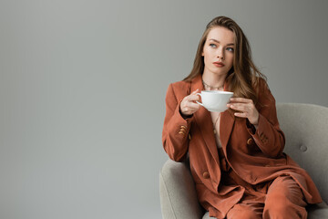 Trendy young and brunette woman in terracotta suit holding cup of cappuccino and looking away while sitting on modern armchair isolated on grey with copy space