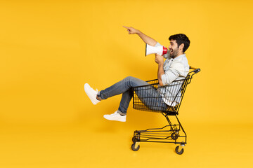 Young man sitting inside of shopping trolley and holding megaphone  isolated on yellow background, Announce concept - obrazy, fototapety, plakaty