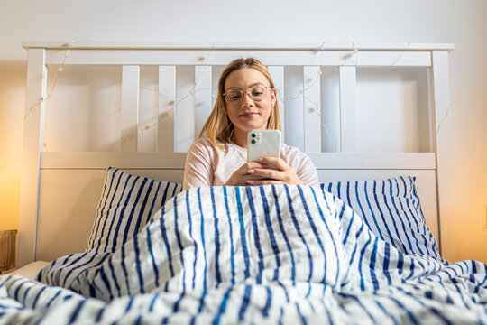 Smiling woman uses her mobile phone in bed in the morning.