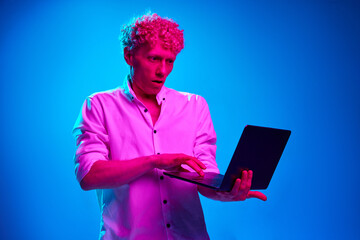 Portrait of curly man in white shirt working with laptop against blue studio background in pink neon light. Shocked face. Concept of human emotions, lifestyle, business, facial expression