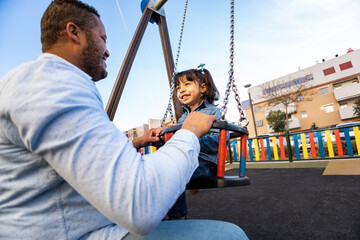 A dark-skinned father happily plays with his daughter on an outdoor playground. The father pushes...