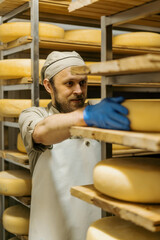 cheese maker in uniform at cheese production man stands in warehouse with wooden shelves full of...