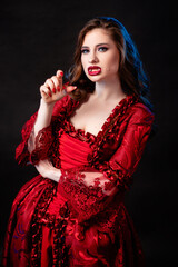 Naklejka premium Portrait of a young, attractive vampire woman in a red rococo dress posing isolated against a dark background with blue backlights.