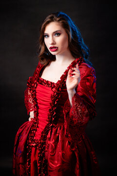 Portrait of a young, attractive vampire woman in a red rococo dress posing isolated against a dark background with blue backlights.