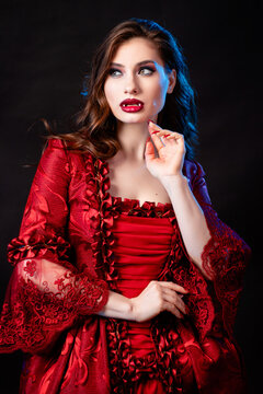 Portrait of a young, attractive vampire woman in a red rococo dress posing isolated against a dark background with blue backlights.