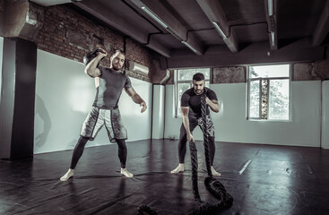 Functional training. Two male athletes are training with a kettlebell and battle ropes in a sports hall