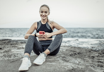 Fit woman in sportswear using smartphone while sitting on urban beach