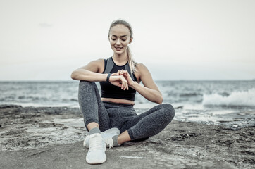 Tired but smiling fit woman in sportswear sitting on urban beach and look at smart bracelet