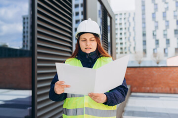 Architect with paper folder with documents at a construction site. Woman constructor wearing helmet and safety yellow vest. Women are planning new building project. Modern exterior