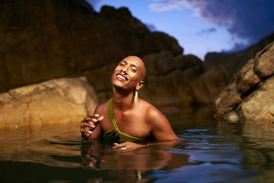 Smiling gender fluid black person poses in natural still water pool. Queer ethnic fashion model in open dress, brass jewelry with gems standing gracefully in the middle of rocky lake at night.