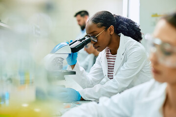 Black female scientist looking through microscope while working in laboratory. - obrazy, fototapety, plakaty