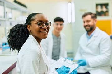 Happy black biochemist working with her colleagues in lab and looking at camera.