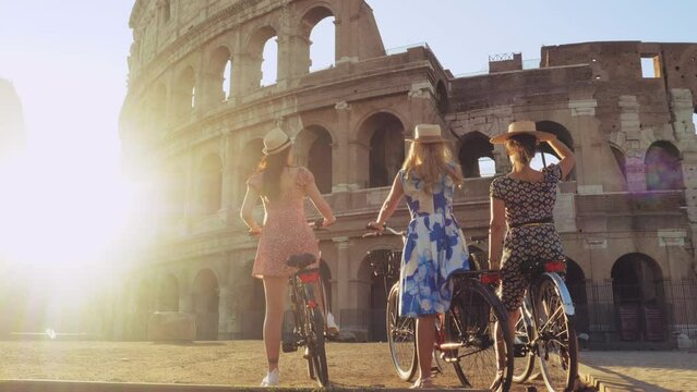 Three happy young women friends tourists with bikes waving hats at Colosseum in Rome, Italy at sunrise.