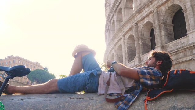 Happy young man tourist lying on a column with bike taking pictures with vintage camera at colosseum in Rome, Italy at sunrise.