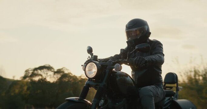Man in a black leather jacket driving a black motorcycle through the dusk sky.