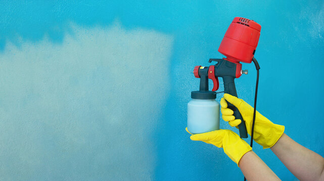 A worker's hands in yellow gloves hold a hand-held spray gun with an overhead compressor. Painting the wall of the house in blue. Household renovation. Copy space. Staining. Airless spraying device