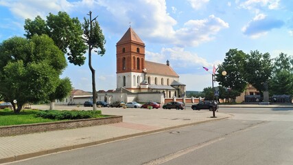 The historic church building with a red brick tower and tile roof is surrounded by a stone fence. Next to it are trees and cars parked in the street. Sunny weather and blue skies with clouds - Powered by Adobe