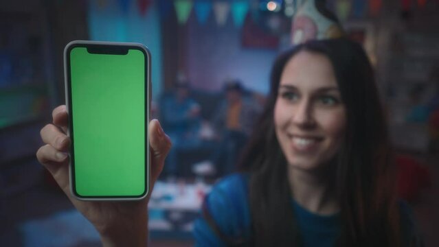 A girl shows a smartphone with a green screen, smiles and nods her head. Close up of a girl with a smartphone on a blurred background of a party, in a room decorated with a disco ball and paper flags.